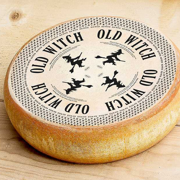 A round cheese wheel with a rustic golden-brown rind and a cream-colored top featuring the words 'OLD WITCH' repeated around the edge and four black silhouettes of witches flying on broomsticks, shown on a light wood surface.