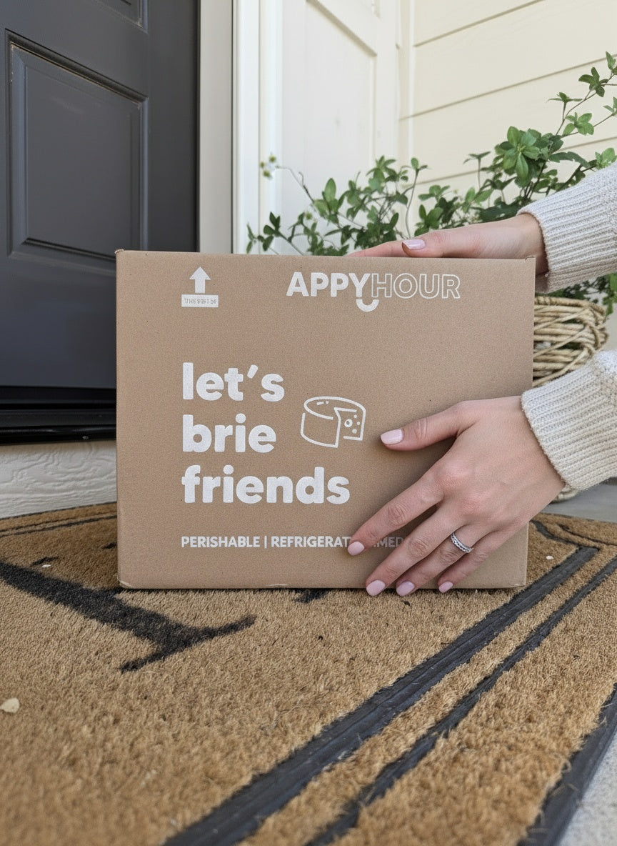 Person holding a 'appy hour' box with 'let's brie friends' text on a doormat.